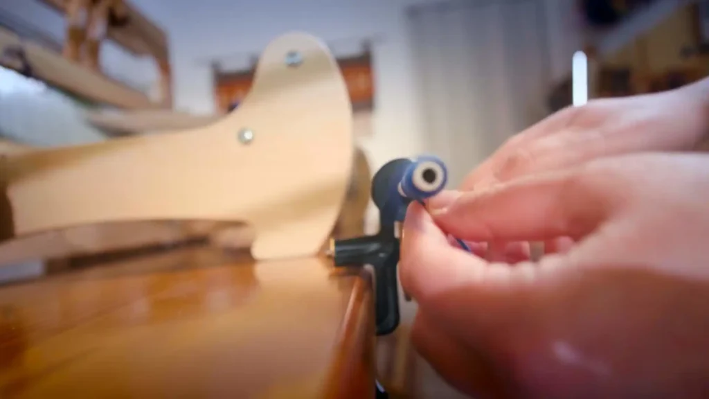Κοντινό πλάνο στα χέρια της Ζωής να τυλίγουν προσεκτικά ένα μπλε νήμα σε ένα μασουράκι χρησιμοποιώντας μια χειροκίνητη μασουρίστρα, με έναν ξύλινο αργαλειό στο φόντο.