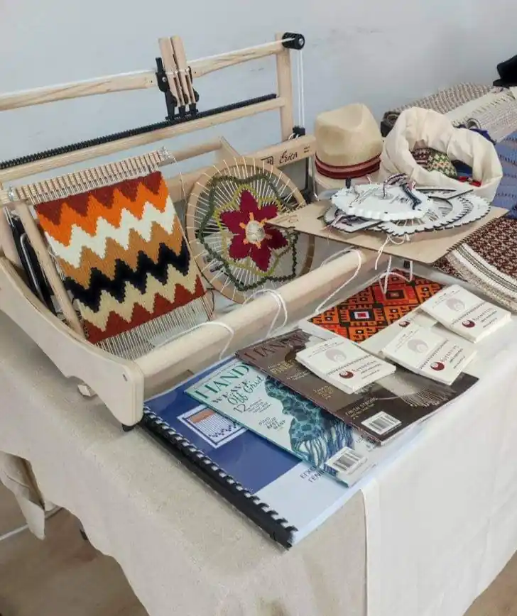 Ένας επιτραπέζιος αργαλειός και πάνω του ένας μικρό τελάρο υφαντικής με υφαντό με γεωμετρικά μοτίβα. Μπροστά βρίσκονται βιβλία υφαντικής.