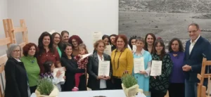 Αναμνηστική ομαδική φωτογραφία από την τελετή αποφοίτησης της Δομής Υφαντικής Ανωγείων. Διακρίνονται οι χαμογελαστές μαθήτριες που αποφοίτησαν κρατώντας τα πιστοποιητικά τους. Στα δεξιά της φωτογραφίας στέκεται ο δήμαρχος Ανωγείων κ. Σωκράτης Κεφαλογιάννης. Στο κέντρο βρίσκεται η εκπαιδεύτρια κ. Ζωή Παπαδάκη, η οποία φοράει κίτρινη μπλούζα, και στα αριστερά διακρίνεται η γραμματέας της δομής κ. Αθηνά, με μαύρο σακάκι. Στον χώρο διακρίνονται ξύλινοι αργαλειοί και μια μεγάλη ασπρόμαυρη φωτογραφία τοπίου στον τοίχο.