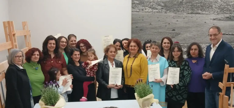 Αναμνηστική ομαδική φωτογραφία από την τελετή αποφοίτησης της Δομής Υφαντικής Ανωγείων. Διακρίνονται οι χαμογελαστές μαθήτριες που αποφοίτησαν κρατώντας τα πιστοποιητικά τους. Στα δεξιά της φωτογραφίας στέκεται ο δήμαρχος Ανωγείων κ. Σωκράτης Κεφαλογιάννης. Στο κέντρο βρίσκεται η εκπαιδεύτρια κ. Ζωή Παπαδάκη, η οποία φοράει κίτρινη μπλούζα, και στα αριστερά διακρίνεται η γραμματέας της δομής κ. Αθηνά, με μαύρο σακάκι. Στον χώρο διακρίνονται ξύλινοι αργαλειοί και μια μεγάλη ασπρόμαυρη φωτογραφία τοπίου στον τοίχο.