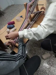 Δύο άτομα στο σεμινάριο υφαντικής συνεργάζονται σε έναν ξύλινο αργαλειό ιμάντα (inkle loom) που βρίσκεται πάνω σε ένα τραπέζι. Δημιουργούν ένα ριγωτό σχέδιο με λευκά, μωβ-φούξια και σκούρα νήματα. Δίπλα στον αργαλειό διακρίνονται κουβάρια νήματος στα αντίστοιχα χρώματα, καθώς και ένα ψαλίδι.