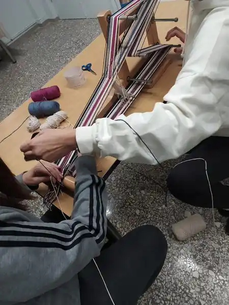 Δύο άτομα στο σεμινάριο υφαντικής συνεργάζονται σε έναν ξύλινο αργαλειό ιμάντα (inkle loom) που βρίσκεται πάνω σε ένα τραπέζι. Δημιουργούν ένα ριγωτό σχέδιο με λευκά, μωβ-φούξια και σκούρα νήματα. Δίπλα στον αργαλειό διακρίνονται κουβάρια νήματος στα αντίστοιχα χρώματα, καθώς και ένα ψαλίδι.