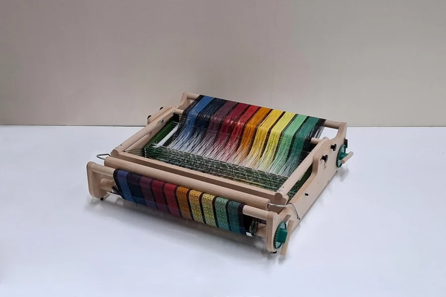 Lojan Flex 8 μιτάρια διπλωμένος Μια λεπτομερής φωτογραφία προϊόντος του επιτραπέζιου αργαλειού 8 μιταριών Lojan Flex πλήρως διπλωμένου flat για εξοικονόμηση χώρου κατά την αποθήκευση και τη μεταφορά. Το πολύχρωμο ριγωτό στημόνι ουράνιου τόξου παραμένει ανέπαφο και υπό τάση, και το ημιτελές υφασμένο ύφασμα είναι ασφαλώς τυλιγμένο, αναδεικνύοντας την ικανότητα του αργαλειού να διπλώνει χωρίς να διαταράσσεται η πρόοδος της ύφανσης. Το προφίλ είναι συμπαγές και οριζόντιο.