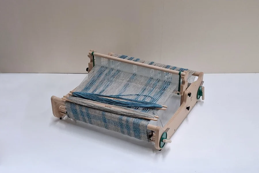 Lojan Flex σταθερού μίτου Μια λεπτομερής φωτογραφία του αργαλειού σταθερού μίτου (rigid heddle) Lojan Flex, πλήρως στημένου με πολύχρωμα μπλε και άσπρα νήματα. Πολλές σαΐτες τυλιγμένες με νήμα πάνω σε ένα ημιτελές υφαντό που εμφανίζει ένα γαλάζιο μοτίβο ορατό από κάτω.