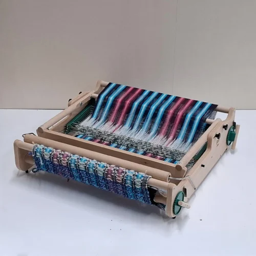 Μια φωτογραφία του επιτραπέζιου αργαλειού 4 μιταριών Lojan Flex, πλήρως διπλωμένου (flat) για εύκολη μεταφορά και εξοικονόμηση χώρου. Ο αργαλειός παραμένει στημένος, με τα πολύχρωμα ριγωτά νήματα και το υφασμένο τμήμα απόλυτα ασφαλή, αναδεικνύοντας πώς διπλώνει χωρίς να επηρεάζεται καθόλου η πρόοδος της ύφανσης.