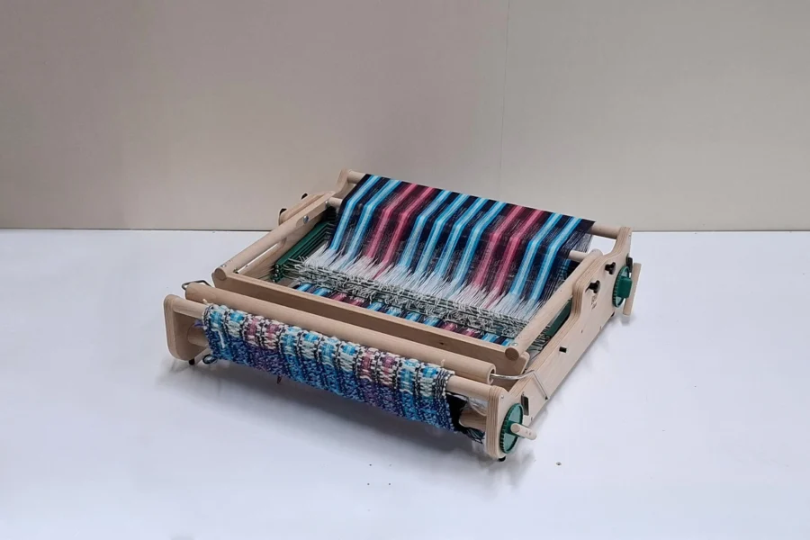 Lojan Flex 4 μιτάρια διπλωμένος Μια φωτογραφία του επιτραπέζιου αργαλειού 4 μιταριών Lojan Flex, πλήρως διπλωμένου (flat) για εύκολη μεταφορά και εξοικονόμηση χώρου. Ο αργαλειός παραμένει στημένος, με τα πολύχρωμα ριγωτά νήματα και το υφασμένο τμήμα απόλυτα ασφαλή, αναδεικνύοντας πώς διπλώνει χωρίς να επηρεάζεται καθόλου η πρόοδος της ύφανσης.