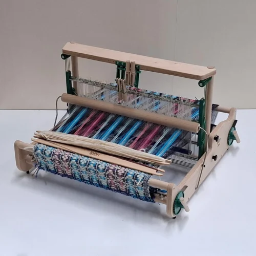 Μια λεπτομερής φωτογραφία του επιτραπέζιου αργαλειού Lojan Flex στη διαμόρφωση με 4 μιτάρια, σε θέση εργασίας. Ο αργαλειός είναι πλήρως στημένος με πολύχρωμα ριγωτά νήματα σε μπλε, ροζ και μοβ αποχρώσεις. Δύο ξύλινες σαΐτες ακουμπούν πάνω στο ημιτελές ύφασμα στο μπροστινό μέρος.