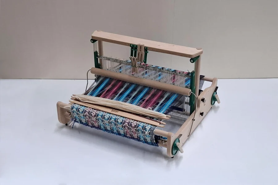 Lojan Flex 4 μιτάρια Μια λεπτομερής φωτογραφία του επιτραπέζιου αργαλειού Lojan Flex στη διαμόρφωση με 4 μιτάρια, σε θέση εργασίας. Ο αργαλειός είναι πλήρως στημένος με πολύχρωμα ριγωτά νήματα σε μπλε, ροζ και μοβ αποχρώσεις. Δύο ξύλινες σαΐτες ακουμπούν πάνω στο ημιτελές ύφασμα στο μπροστινό μέρος.