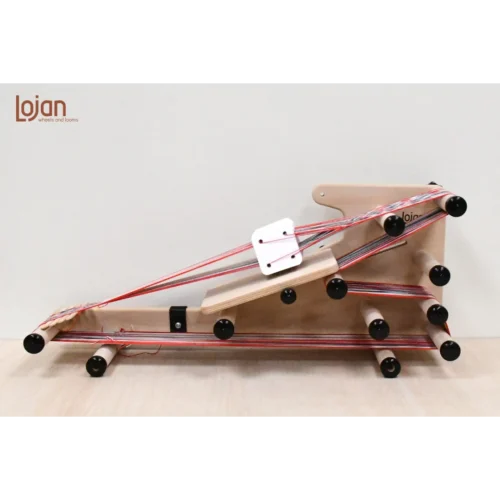 Μια λεπτομερής πλάγια όψη του αργαλειού Inkle 3-1 της Lojan με βοηθητική πλάκα στήριξης για υφαντική με κάρτες. Ο αργαλειός είναι στημένος με πολύχρωμα νήματα με κόκκινες και γκρι ρίγες. Λευκές τετράγωνες κάρτες ύφανσης είναι περασμένες στο στημόνι.