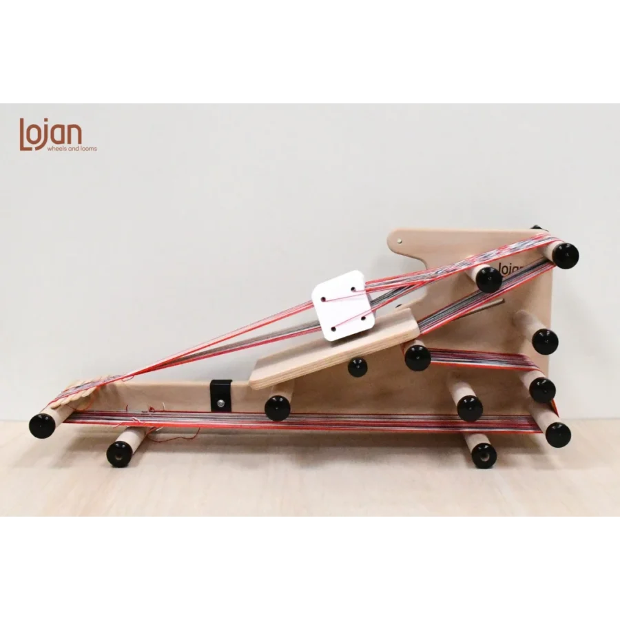 Μια λεπτομερής πλάγια όψη του αργαλειού Inkle 3-1 της Lojan με βοηθητική πλάκα στήριξης για υφαντική με κάρτες. Ο αργαλειός είναι στημένος με πολύχρωμα νήματα με κόκκινες και γκρι ρίγες. Λευκές τετράγωνες κάρτες ύφανσης είναι περασμένες στο στημόνι.