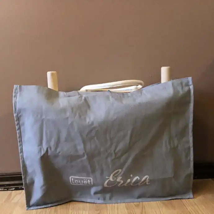 Γκρι υφασμάτινη τσάντα μεταφοράς τύπου tote για τον αργαλειό Louët Erica, ακουμπισμένη σε πάτωμα. Στο κάτω μέρος της τσάντας διακρίνεται το λογότυπο της Louët και η λέξη Erica. Από το πάνω ανοιχτό μέρος προεξέχουν ελαφρώς οι ξύλινες άκρες του διπλωμένου αργαλειού και τα λευκά χερούλια.