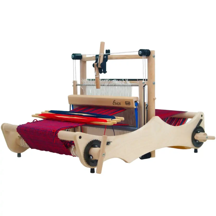 Louet Erica με 2 μιτάρια Ο επιτραπέζιος αργαλειός Louët Erica με 2 μιτάρια, ανοιχτός και έτοιμος για ύφανση. Ο αργαλειός είναι στημένος με στημόνι σε αποχρώσεις του κόκκινου και του μπλε, και έχει μερικώς υφασμένο ύφασμα. Πάνω στο ύφασμα ακουμπούν δύο ξύλινες σαΐτες με κόκκινο και μπλε νήμα.