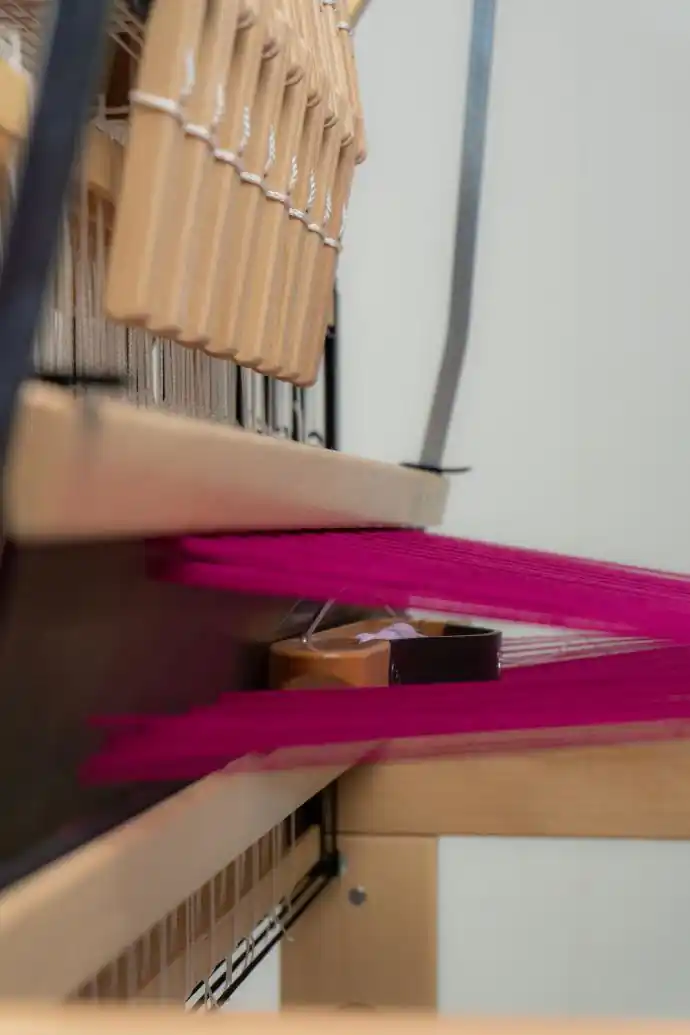 Μια κοντινή, υπό γωνία άποψη του αργαλειού Louet Jane, που αναδεικνύει το εξαιρετικά μεγάλο και καθαρό διάκενο του στημονιού. Το έντονο φούξια στημόνι είναι χωρισμένο, δημιουργώντας ένα ευρύχωρο άνοιγμα. Μια ξύλινη σαΐτα βάρκα με μασούρι διακρίνεται βαθιά μέσα στο διάκενο, επιδεικνύοντας πόσο άνετα και εύκολα μπορεί να περάσει.