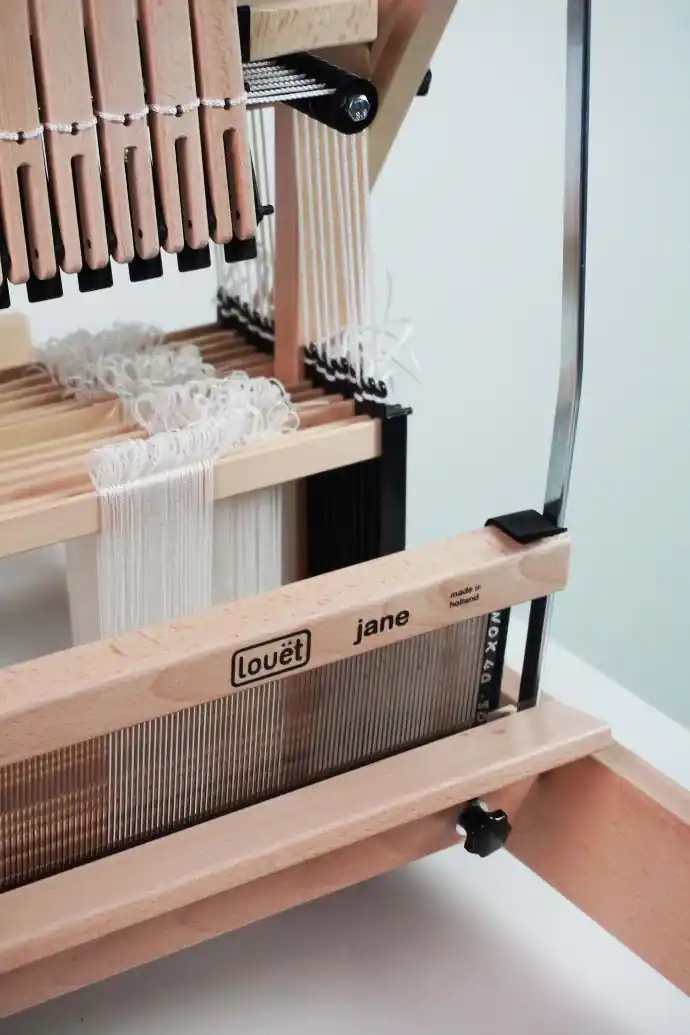 Κοντινή άποψη του επιτραπέζιου αργαλειού Louet Jane, εστιάζοντας στην κάτω μπάρα στήθους με τα λογότυπα "louët" και "jane" και την επιγραφή "made in holland". Ακριβώς κάτω από τη μπάρα είναι εγκατεστημένο το μεταλλικό χτένι. Διακρίνονται επίσης τα μιτάρια με τα λευκά νήματα και μέρος του μηχανισμού των μιταριών με τους ξύλινους μοχλούς.
