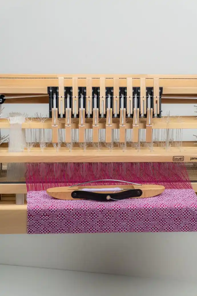 Μια κοντινή φωτογραφία από μπροστά του επιτραπέζιου αργαλειού Louet Jane, που εστιάζει στους μοχλούς χειρισμού των 8 μιταριών. Διακρίνεται ένα στημόνι έντονου φούξια χρώματος και μια σαΐτα βάρκα με μασούρι, η οποία ακουμπά πάνω σε ένα ημιτελές υφαντό ύφασμα με μωβ και ροζ γεωμετρικό μοτίβο. Τα λογότυπα Louet και Jane είναι ορατά.