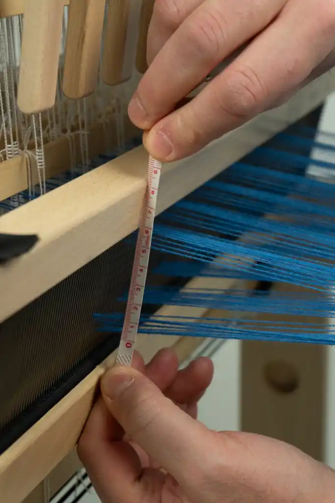 Μια κοντινή φωτογραφία ενός χεριού που χρησιμοποιεί ένα μικρό χάρακα για να μετρήσει το ύψος του ανοίγματος του στημονιού στο χτένι του αργαλειού Jane. Μπλε νήματα στημονιού περνούν μέσα από το χτένι. Το χέρι κρατά το χάρακα σταθερά, δείχνοντας τη μέτρηση (περίπου 5-6 εκ).