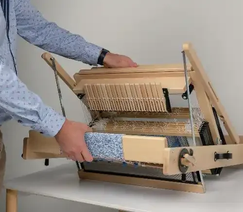 Ένας άνδρας διπλώνει τον επιτραπέζιο αργαλειό Louët Jane. Η φωτογραφία εστιάζει στα χέρια του που κρατούν το ξύλινο πλαίσιο, επιδεικνύοντας πώς ο αργαλειός 8 μιταριών αναδιπλώνεται σε ένα συμπαγές σχήμα, ακόμα και με το υφαντό πάνω του.