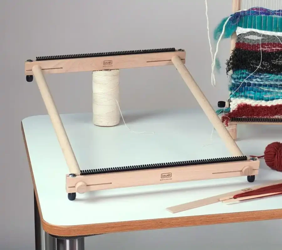 Louet Lisa Medium - WP0157 Το μεσαίο μέγεθος του ξύλινου τελάρου υφαντικής Louët Lisa, άδειο και συναρμολογημένο πάνω σε ένα τραπέζι. Οι μπάρες στημονιού φέρουν το λογότυπο της Louët. Ένας μεγάλος κώνος με λευκό νήμα στημονιού βρίσκεται όρθιος μέσα στο πλαίσιο. Στο βάθος, στα δεξιά, διακρίνεται ένα άλλο τελάρο με ημιτελή, πολύχρωμη ταπετσαρία.