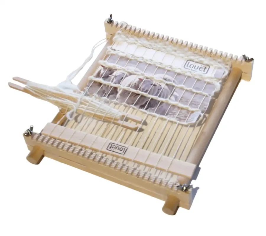 Louet Lisa Small - WP0156 Ο μικρός ξύλινος αργαλειός-τελάρο Louet Lisa, πλήρως συναρμολογημένος και στημένος με λευκό στημόνι. Μια ξύλινη σαΐτα με λευκό νήμα ακουμπά πάνω στα νήματα. Πίσω από το στημόνι έχει τοποθετηθεί μια κάρτα με φωτογραφία από ένα κοπάδι πρόβατα και το λογότυπο της Louët, η οποία χρησιμεύει ως οπτικός οδηγός/πατρόν για την ύφανση της ταπετσαρίας.