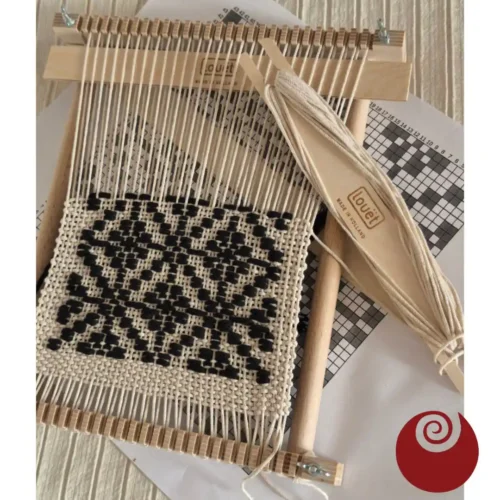 Louet Lisa υφαντική της Ζωής Φωτογραφία από πάνω του μικρού ξύλινου τελάρου υφαντικής Louët Lisa. Πάνω στο τελάρο βρίσκεται ένα όμορφο ασπρόμαυρο γεωμετρικό υφαντό, δημιουργία της Ζωής. Μια επίπεδη ξύλινη σαΐτα της Louët, τυλιγμένη με λευκό νήμα, ακουμπά διαγώνια πάνω στο έργο. Κάτω από το τελάρο διακρίνεται το τυπωμένο χάρτινο πατρόν με το σχέδιο της ύφανσης. Στην κάτω δεξιά γωνία φαίνεται το κόκκινο σπειροειδές λογότυπο της Εργαστίνης.