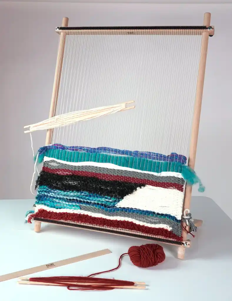 Louet Lisa XL - WP0159 Το πολύ μεγάλο (XL) μέγεθος του ξύλινου τελάρου υφαντικής Louët Lisa, όρθιο πάνω σε τραπέζι. Είναι στημένο και διαθέτει μια εντυπωσιακή, παχιά ταπισερί ελεύθερης ύφανσης σε αποχρώσεις του τιρκουάζ, κόκκινου, γκρι, μαύρου και λευκού. Μια σαΐτα με λευκό νήμα κρέμεται από το στημόνι. Μπροστά από το τελάρο υπάρχουν εργαλεία υφαντικής Louët και ένα κουβάρι μπορντό νήμα.