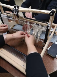 Κοντινό πλάνο από εργαστήριο υφαντικής σε επιτραπέζιο αργαλειό. Φαίνονται τα χέρια ενός ατόμου καθώς προετοιμάζει τον ξύλινο αργαλειό (μίτωμα), χρησιμοποιώντας έναν κόκκινο κλέφτη. Στο βάθος διακρίνονται τα χέρια ενός δεύτερου ατόμου, καθώς το μίτωμα συνήθως απαιτεί δύο άτομα.