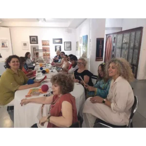 Ομάδα γυναικών σε εργαστήριο υφαντικής στο ΜΕΤΗ. Κάθονται γύρω από ένα στρογγυλό τραπέζι και εργάζονται σε μικρούς, φορητούς αργαλειούς. Η Ζωή κοιτάζει προς την κάμερα