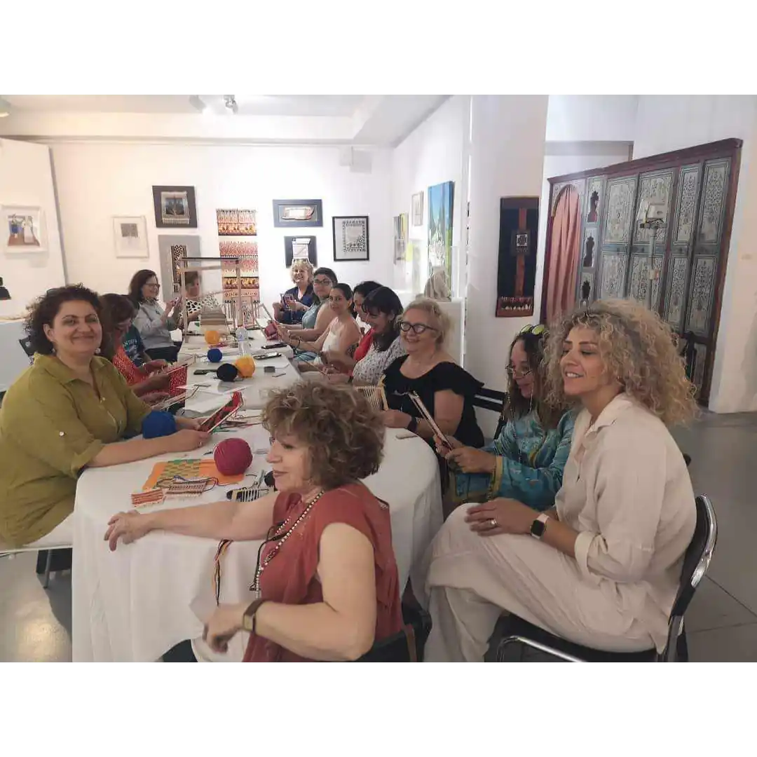 Ομάδα γυναικών σε εργαστήριο υφαντικής στο ΜΕΤΗ. Κάθονται γύρω από ένα στρογγυλό τραπέζι και εργάζονται σε μικρούς, φορητούς αργαλειούς. Η Ζωή κοιτάζει προς την κάμερα