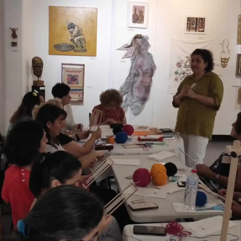 Ενήλικες συμμετέχουσες σε εργαστήριο υφαντικής στο ΜΕΤΗ. Κάθονται σε τραπέζια και εργάζονται σε μικρούς, ατομικούς αργαλειούς. Η Ζωή, με πράσινη μπλούζα, στέκεται και καθοδηγεί.