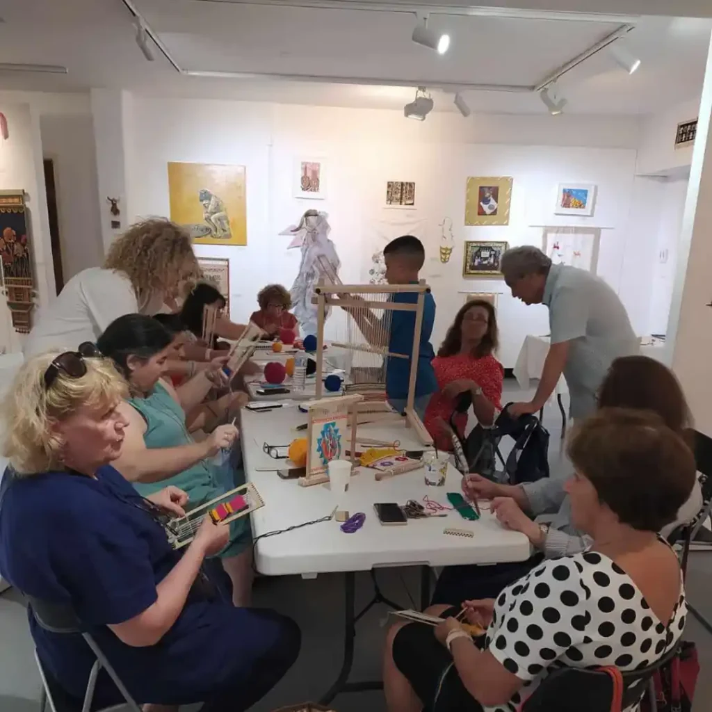 Γενική άποψη του εργαστηρίου υφαντικής στο Μουσείο Εικαστικών Τεχνών Ηρακλείου. Συμμετέχοντες, κυρίως γυναίκες, εργάζονται σε τραπέζια με μικρούς αργαλειούς. Η Ζωή καθοδηγεί. Στο βάθος, η έκθεση «Αρωδαμοί και Ασπάλαθοι».