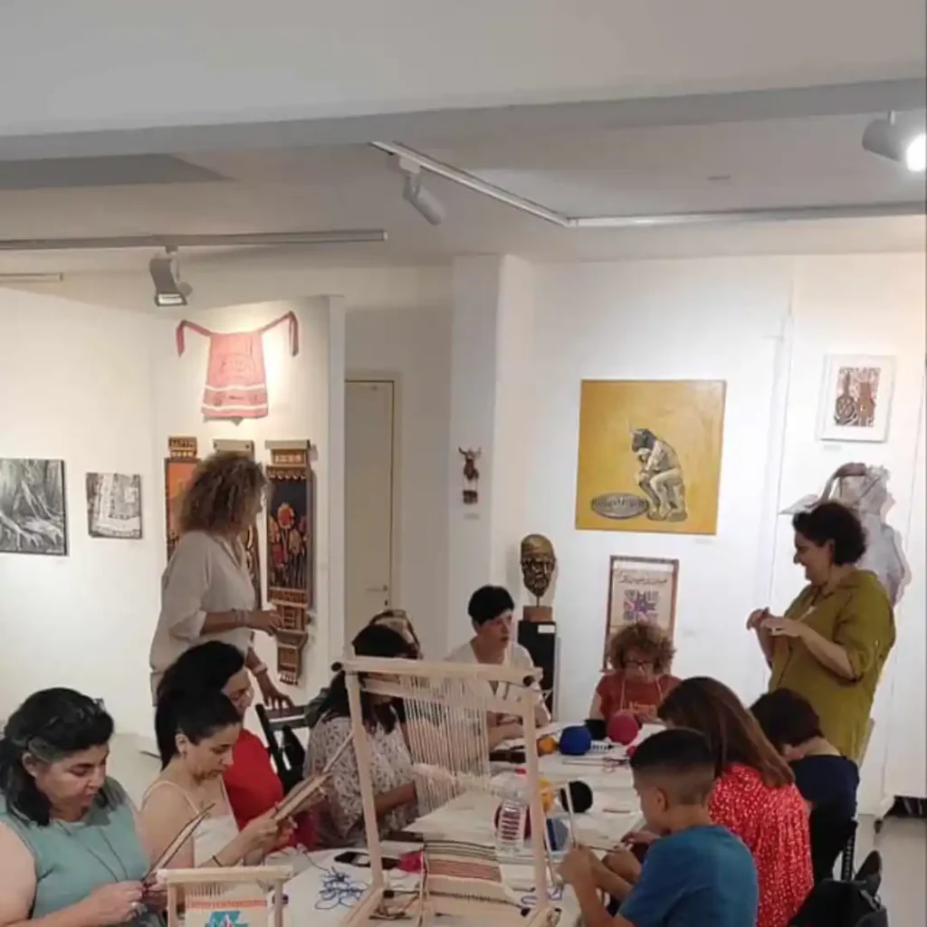Συμμετέχοντες σε εργαστήριο υφαντικής στο Μουσείο Εικαστικών Τεχνών Ηρακλείου, ενήλικες και ένα παιδί, εργάζονται σε μικρούς αργαλειούς. Η Ζωή στέκεται και δείχνει κάτι. Έμφαση στα χέρια και τα νήματα.