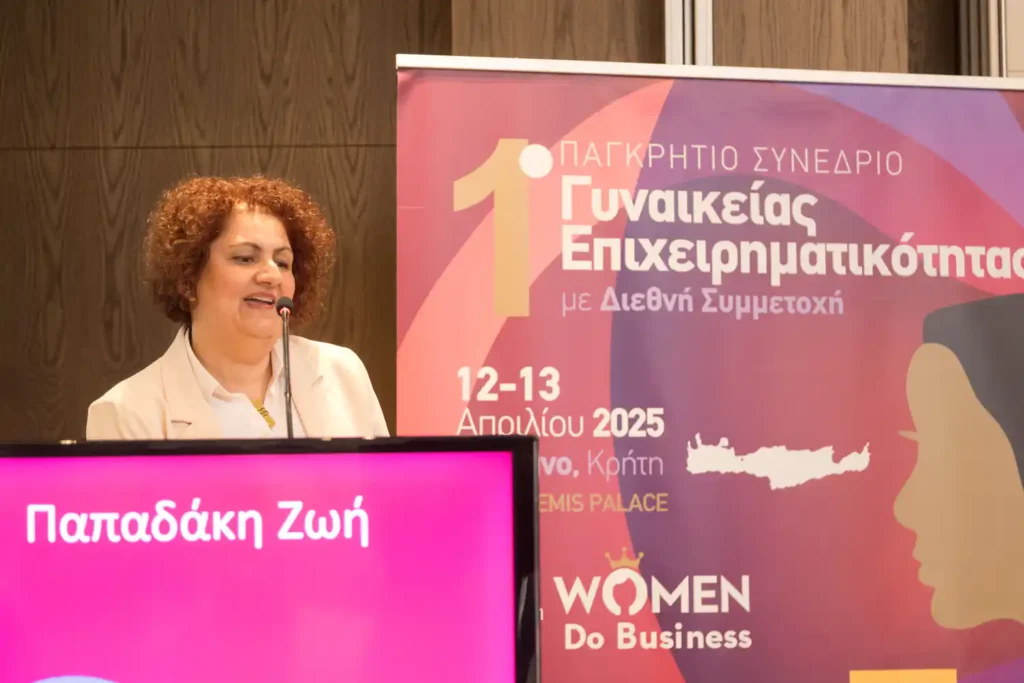 Η Ζωή Παπαδάκη στο βήμα του ομιλητή κατά τη διάρκεια της ομιλίας της στο 1ο Παγκρήτιο Συνέδριο Γυναικείας Επιχειρηματικότητας. Μπροστά της υπάρχει ψηφιακή οθόνη με το όνομά της, ενώ στο φόντο διακρίνεται η κεντρική αφίσα του συνεδρίου από το Women Do Business.