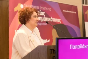 Η Ζωή Παπαδάκη φωτογραφημένη σε προφίλ ενώ μιλάει στο μικρόφωνο από το βήμα του 1ου Παγκρήτιου Συνεδρίου Γυναικείας Επιχειρηματικότητας. Φοράει λευκό σακάκι με μια χρυσή καρφίτσα σε σχήμα σπείρας (το λογότυπο της Εργαστίνης). Πίσω της δεσπόζει το μεγάλο πολύχρωμο banner του συνεδρίου, ενώ στο κάτω μέρος δεξιά διακρίνεται τμήμα μιας οθόνης με το επίθετό της.